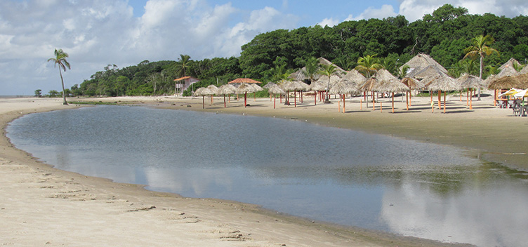 Foto mostra visão panorâmica da praia do Pesqueiro, na Ilha do Marajó. Há uma pequena lagoa formada por maré seca, palhoças vazias e vegetação nativa ao fundo.